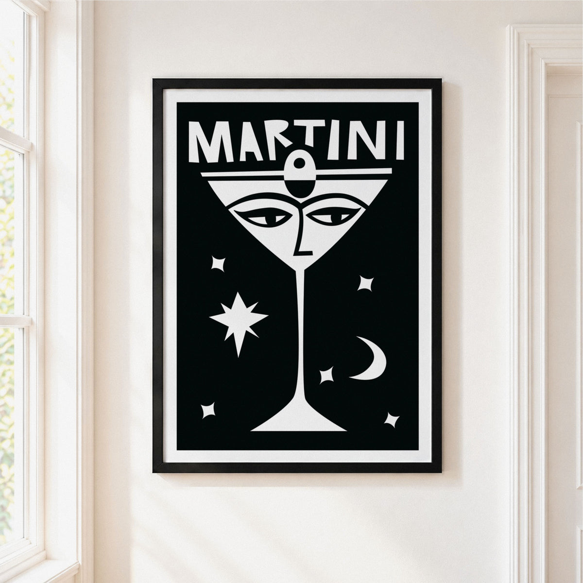 A framed black and white art print featuring a stylized martini glass with a face and the word 'MARTINI' at the top, hanging on a white wall next to a window.