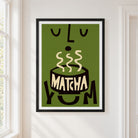 A framed art print featuring a stylized green matcha bowl with cream-colored steam and the word 'MATCHA' in cream, hanging on a white wall next to a window.