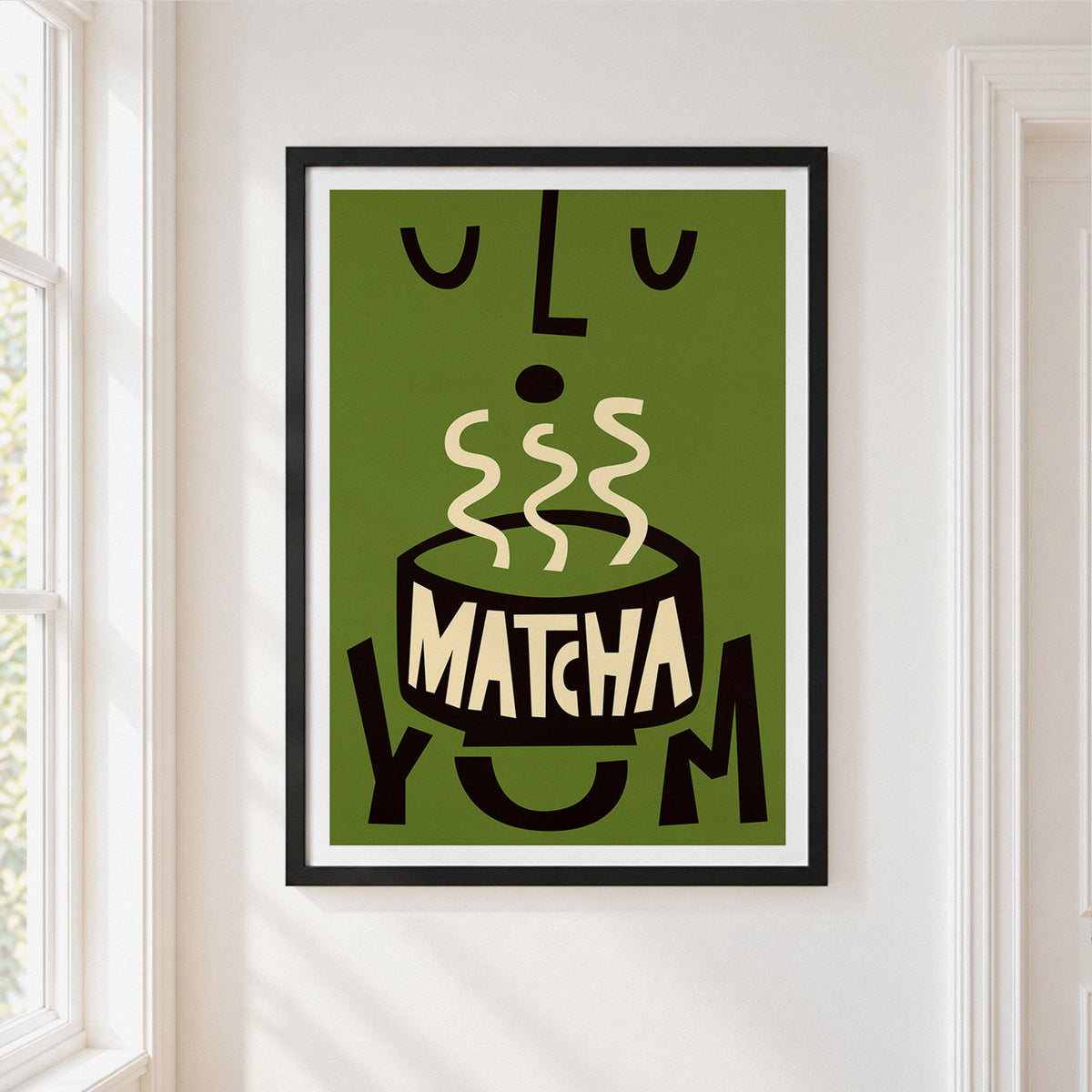 A framed art print featuring a stylized green matcha bowl with cream-colored steam and the word 'MATCHA' in cream, hanging on a white wall next to a window.