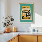 A framed art print featuring a stylized yellow and black egg-shaped design on a teal background, hanging on a white kitchen wall above a counter with a potted plant and mugs.