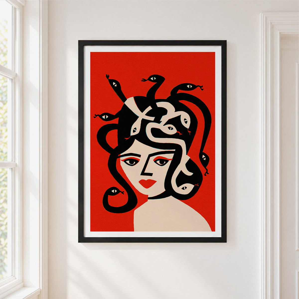 A framed art print featuring a stylized illustration of Medusa with black snakes in her hair, set against a bold red background, hanging on a white wall next to a window.