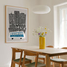 A framed art print featuring a stylized black and white city skyline with a blue and white bus and the word 'MIAMI' in a light wood dining room, styled with a yellow vase of flowers on a wooden table.