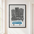 Framed art print of a stylized Miami skyline with black buildings, blue and white bus illustration, and the word 'MIAMI' on a light gray background, hanging on a cream wall.