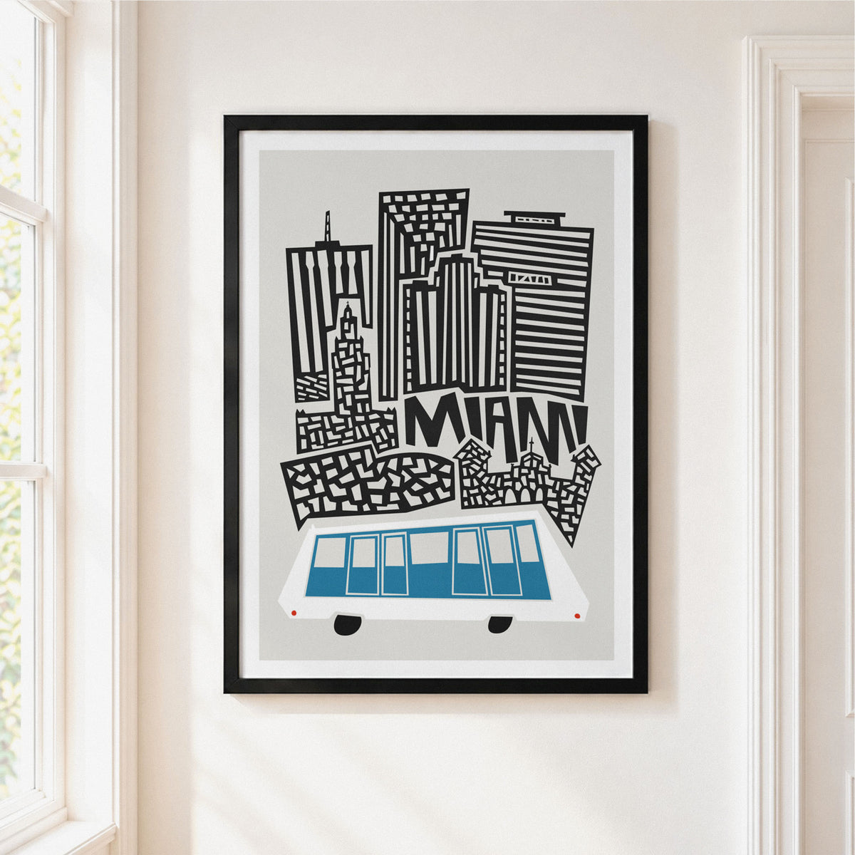 Framed art print of a stylized Miami skyline with black buildings, blue and white bus illustration, and the word 'MIAMI' on a light gray background, hanging on a cream wall.