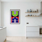 A framed abstract geometric wall art print hangs on a white wall in a modern kitchen setting. The artwork features a split design with green circles on a lavender background on the top half, and blue circles on a red background on the bottom half, enclosed in a thin black frame.
