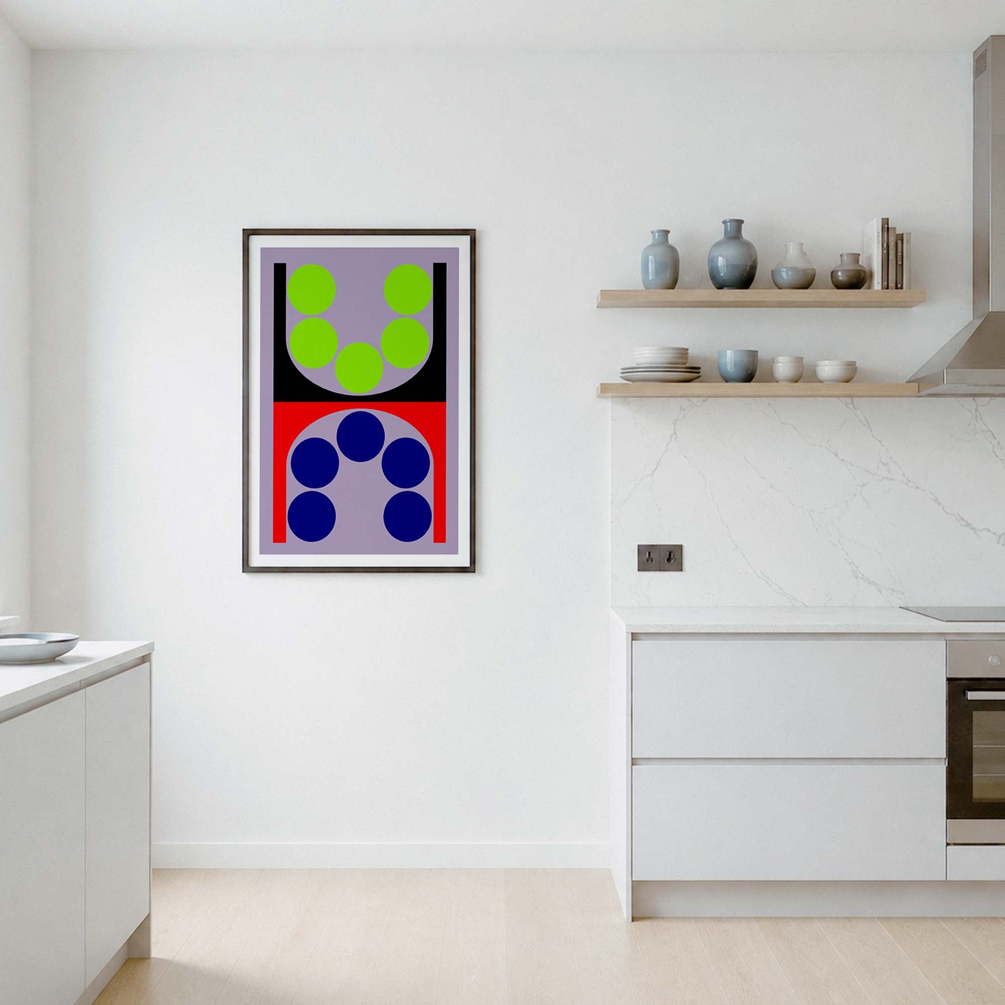 A framed abstract geometric wall art print hangs on a white wall in a modern kitchen setting. The artwork features a split design with green circles on a lavender background on the top half, and blue circles on a red background on the bottom half, enclosed in a thin black frame.