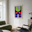 A framed abstract geometric wall art print hangs on a white wall in a modern living room. The artwork features bold shapes in lime green, navy blue, red, black, and lavender.