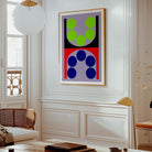 A framed abstract geometric wall art print hangs on a white paneled wall in a bright, modern living room. The artwork features a split design with green circles on a lavender background in the top half, and blue circles on a red background in the bottom half, enclosed in a light wood frame.