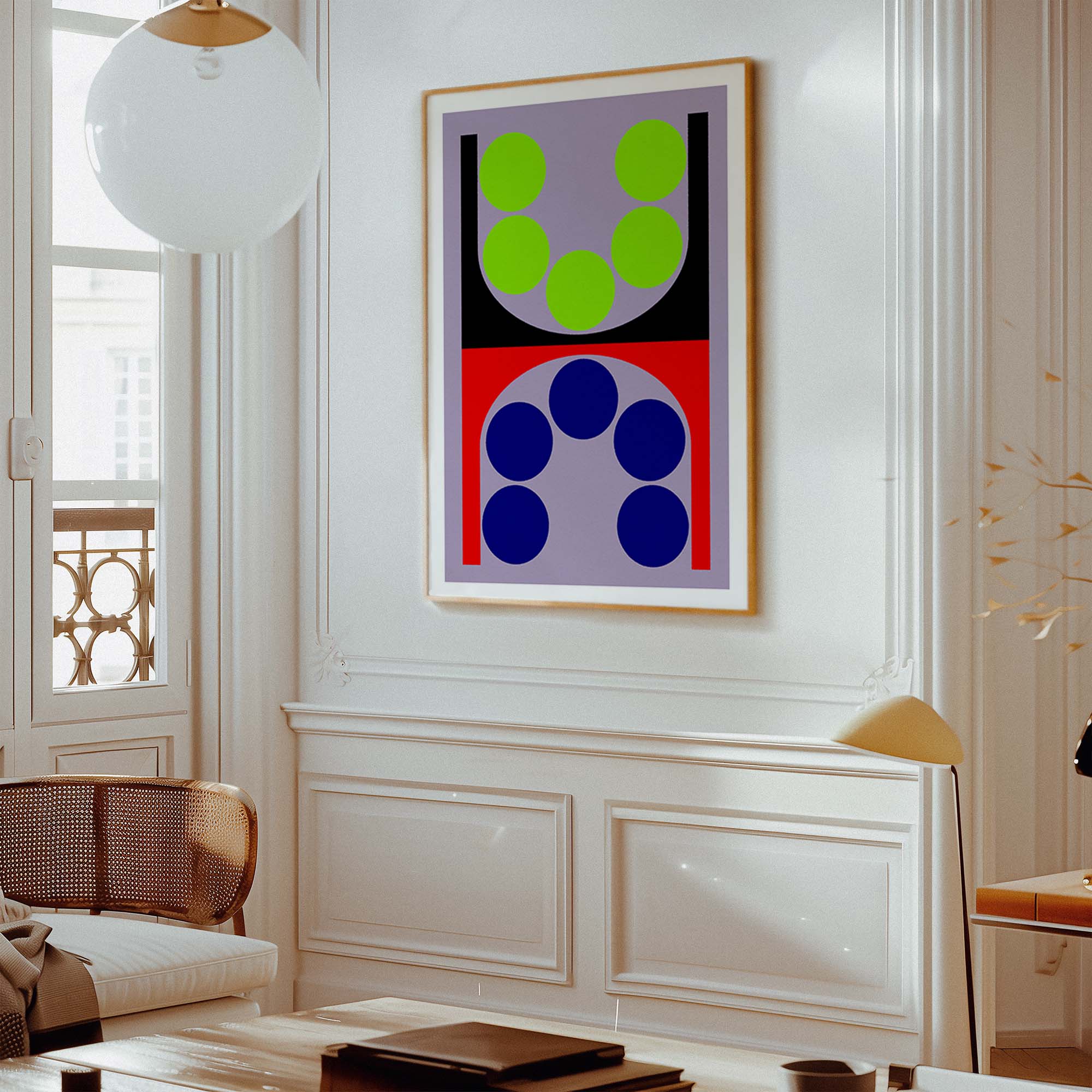 A framed abstract geometric wall art print hangs on a white paneled wall in a bright, modern living room. The artwork features a split design with green circles on a lavender background in the top half, and blue circles on a red background in the bottom half, enclosed in a light wood frame.