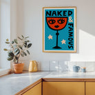 A framed art print featuring a retro-style illustration of a wine glass with a face and the text 'NAKED & FAMOUS' in teal, orange, cream, and black. The print is displayed on a white wall in a modern kitchen setting with a potted plant, yellow pitcher, and mugs on the counter.