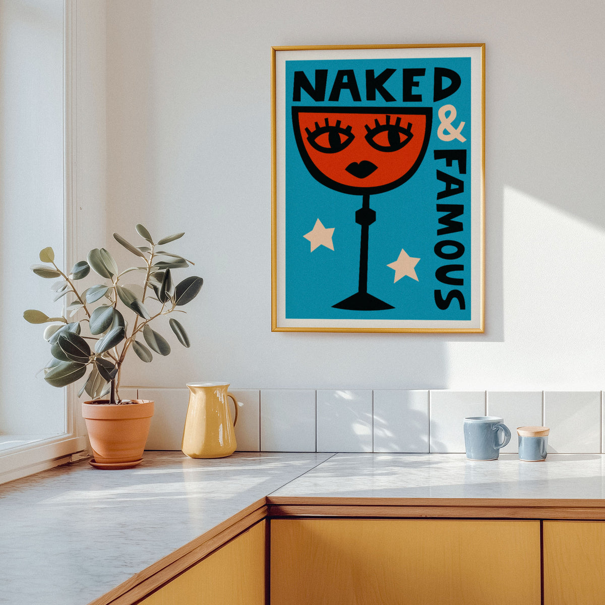 A framed art print featuring a retro-style illustration of a wine glass with a face and the text 'NAKED & FAMOUS' in teal, orange, cream, and black. The print is displayed on a white wall in a modern kitchen setting with a potted plant, yellow pitcher, and mugs on the counter.