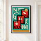 A framed abstract art print featuring the word 'NEGRONI' in cream lettering over red geometric shapes on a teal background, hung on a white wall next to a window.