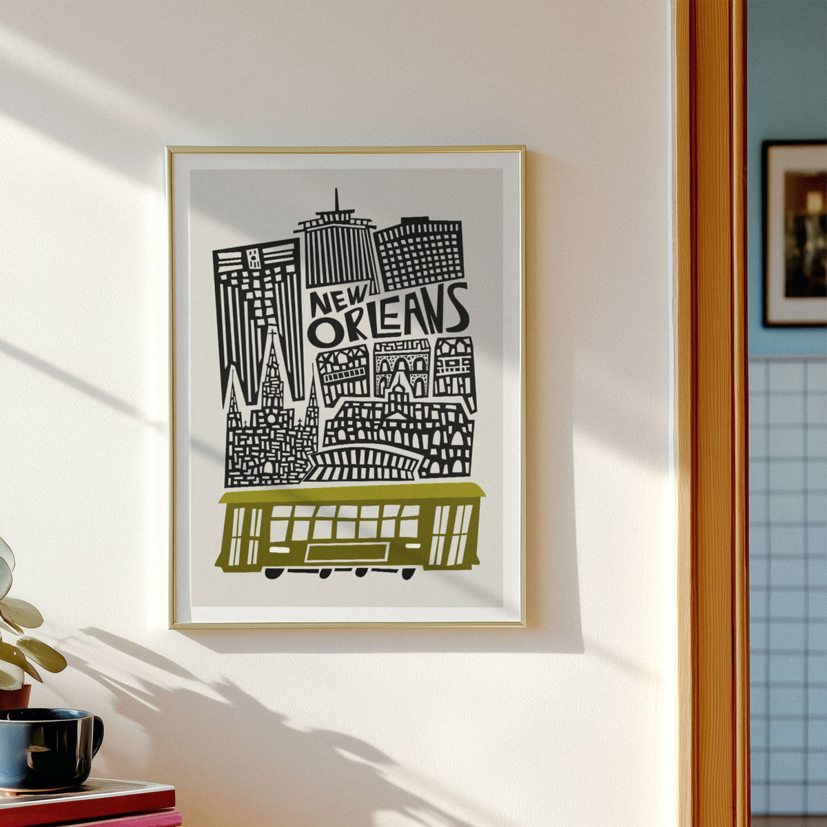A framed New Orleans cityscape art print hangs on a white wall. The print features black architectural silhouettes and the words 'NEW ORLEANS' above a yellow-green streetcar, set in a light wood frame.
