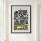 Framed New Orleans cityscape wall art hanging on a cream wall beside a window. The print shows black architectural silhouettes and the words “NEW ORLEANS” with a muted olive-yellow streetcar at the bottom, set in a black frame with a white mat.