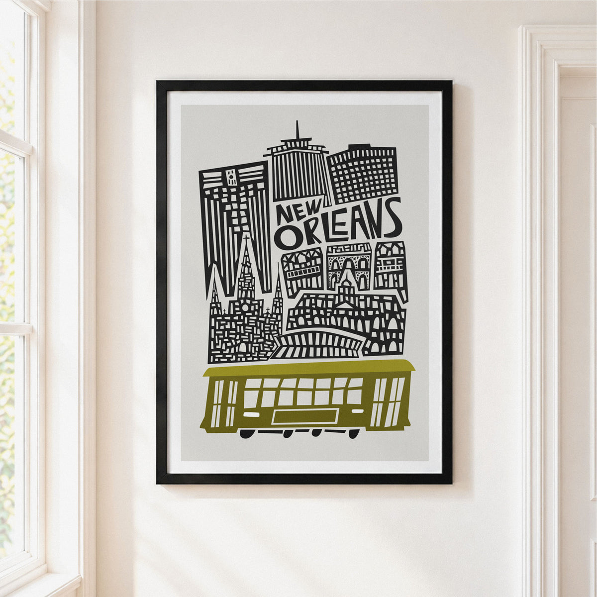 Framed New Orleans cityscape wall art hanging on a cream wall beside a window. The print shows black architectural silhouettes and the words “NEW ORLEANS” with a muted olive-yellow streetcar at the bottom, set in a black frame with a white mat.