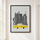 A framed art print featuring a stylized New York skyline with a yellow taxi and black architectural details on a light gray background, hung on a cream-colored wall next to a window.