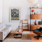 A framed art print featuring a black and white illustration of Newcastle with a yellow train, displayed on a white wall in a modern, sunlit living room with a wooden coffee table, beige sofa, and mid-century modern furniture.