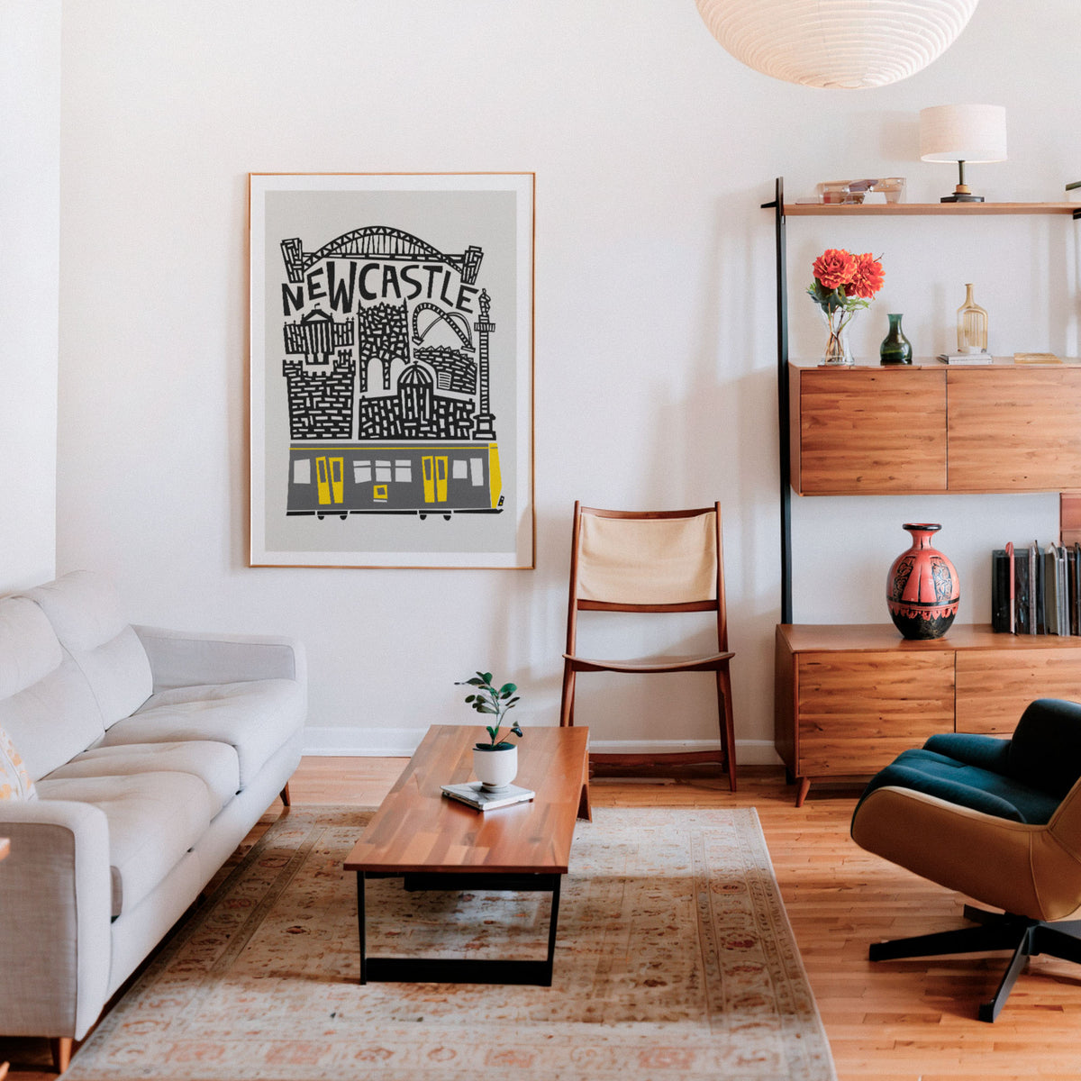 A framed art print featuring a black and white illustration of Newcastle with a yellow train, displayed on a white wall in a modern, sunlit living room with a wooden coffee table, beige sofa, and mid-century modern furniture.