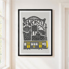 A framed art print featuring a stylized black and gray illustration of Newcastle with a yellow tram, hanging on a white wall in a bright room.