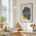 A framed art print featuring a black and white illustration of Norwich cityscape with a bright yellow bird, hanging on a light wall in a modern kitchen setting.