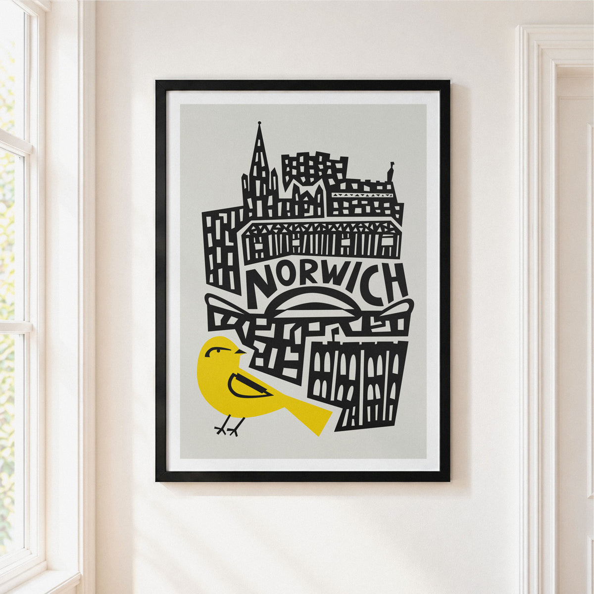 A framed art print featuring a stylized black-and-white illustration of Norwich cityscape with a bright yellow bird, hanging on a white wall next to a window.