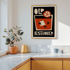 A framed retro-style art print with the words 'OLD FASHIONED' hangs on a white kitchen wall above a countertop. The print features a bold black background, an orange cocktail glass, cream lettering, and a gold frame.