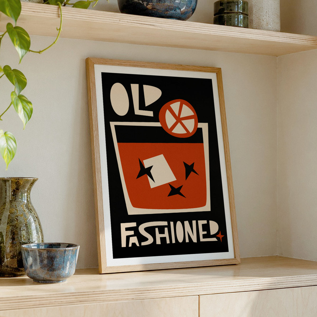 A framed art print leaning on a light wood shelf, featuring bold retro typography and a red-orange cocktail glass illustration on a black background. The print is styled in a warm interior setting with ceramic vases and a trailing green plant nearby.