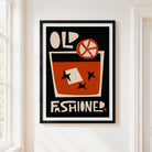 A framed vintage-style poster hanging on a white wall beside a window. The poster features a black background with cream and rust-orange graphic elements, including the words 'OLD FASHIONED' and a stylized drink illustration with a garnish.
