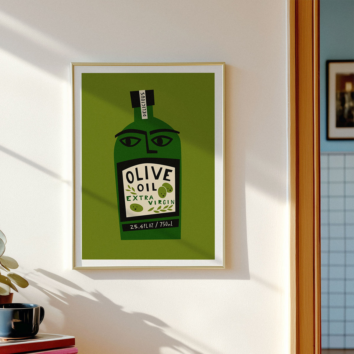 A framed art print featuring a stylized green olive oil bottle with a face, hung on a white wall in a sunlit room. The print reads 'OLIVE OIL EXTRA VIRGIN' and includes a small 'DELICIOUS' mark on the neck label.