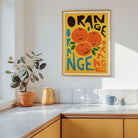 A framed retro-style orange poster hangs on a white kitchen wall above a countertop, with a potted plant, yellow pitcher, and blue mugs nearby.