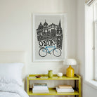 A framed black and white art print featuring a blue bicycle and the word 'OXFORD' hangs on a white wall above a yellow bedside table in a styled bedroom setting.