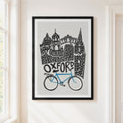 A framed art print featuring a stylized Oxford skyline with a blue bike and black architectural details on a light gray background, hung on a cream-colored wall next to a window.