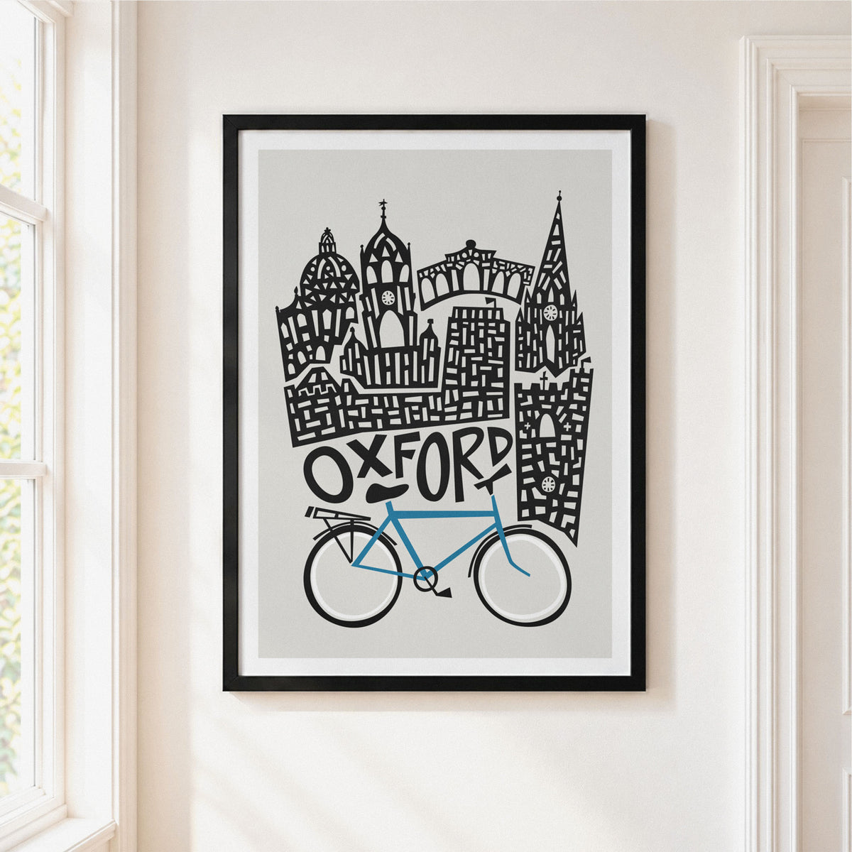 A framed art print featuring a stylized Oxford skyline with a blue bike and black architectural details on a light gray background, hung on a cream-colored wall next to a window.