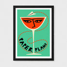 Framed art print featuring a stylized orange cocktail glass with eyes and a paper airplane on a mint green background, with the words 'PAPER PLANE' in black along the stem, shown against a light gray wall.