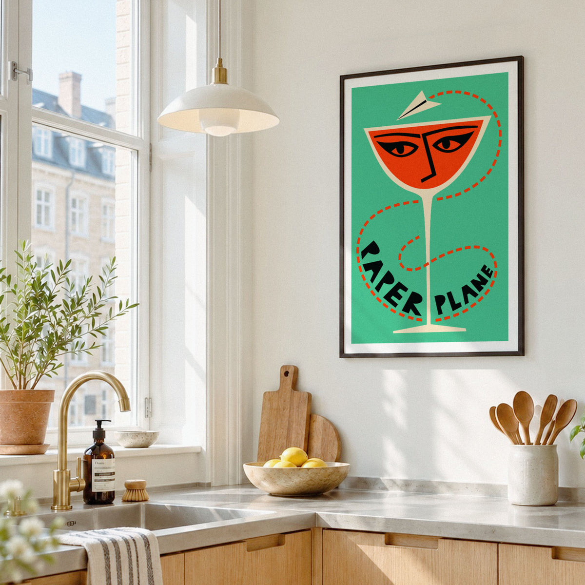 A framed pop-art style poster featuring a yellow mayonnaise can with red, black, and white typography, hanging on a white kitchen wall above a countertop with a sink, plants, and utensils.