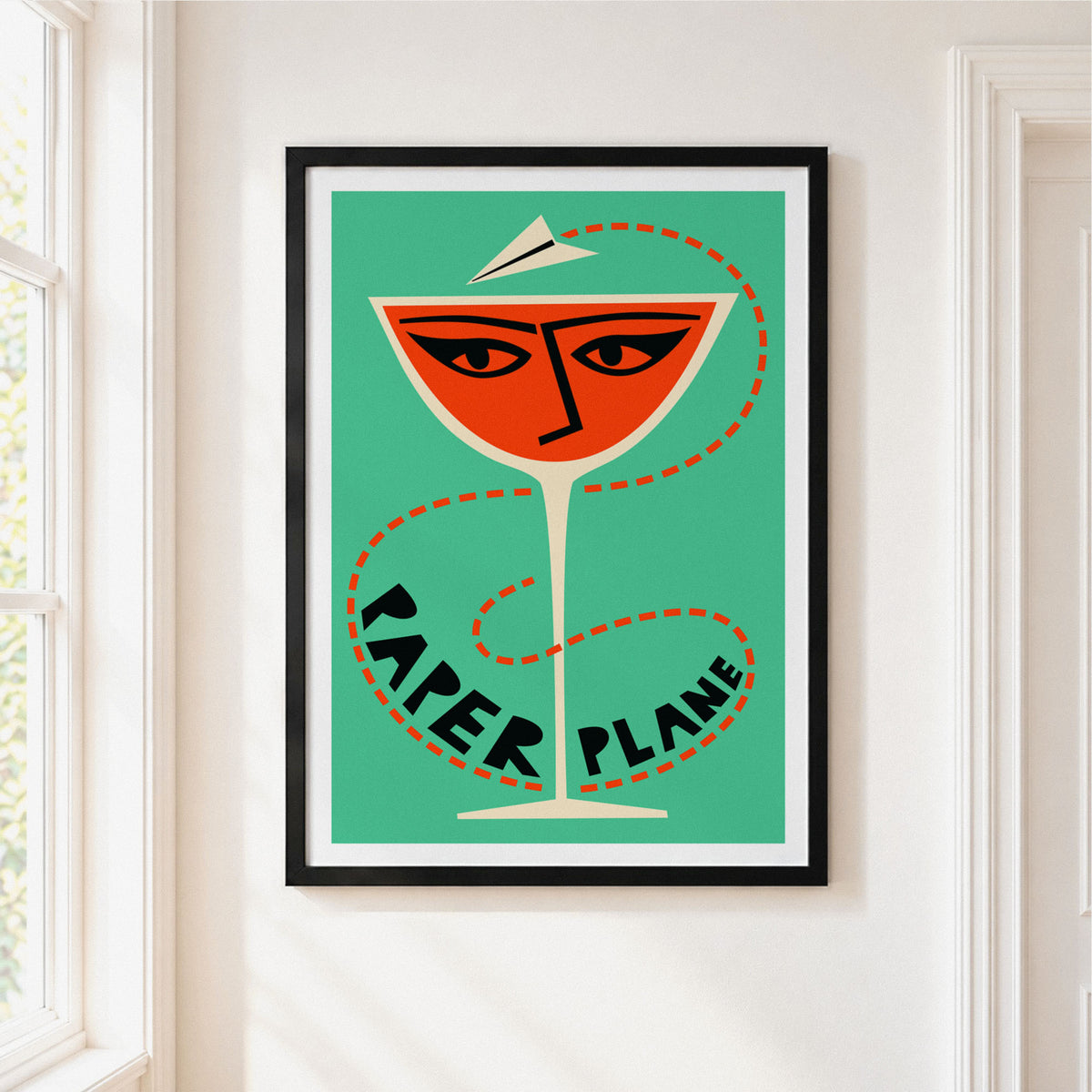 A framed art print featuring a stylized cocktail glass face with eyes and a paper plane hat, on a teal background with the words 'PAPER PLANE' in black. The print is displayed on a white wall next to a window.