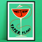 A bold cocktail print featuring a vivid orange drink in a coupe glass, illustrated with a stylised face and topped with a flying paper plane, all set against a mint green backdrop.
