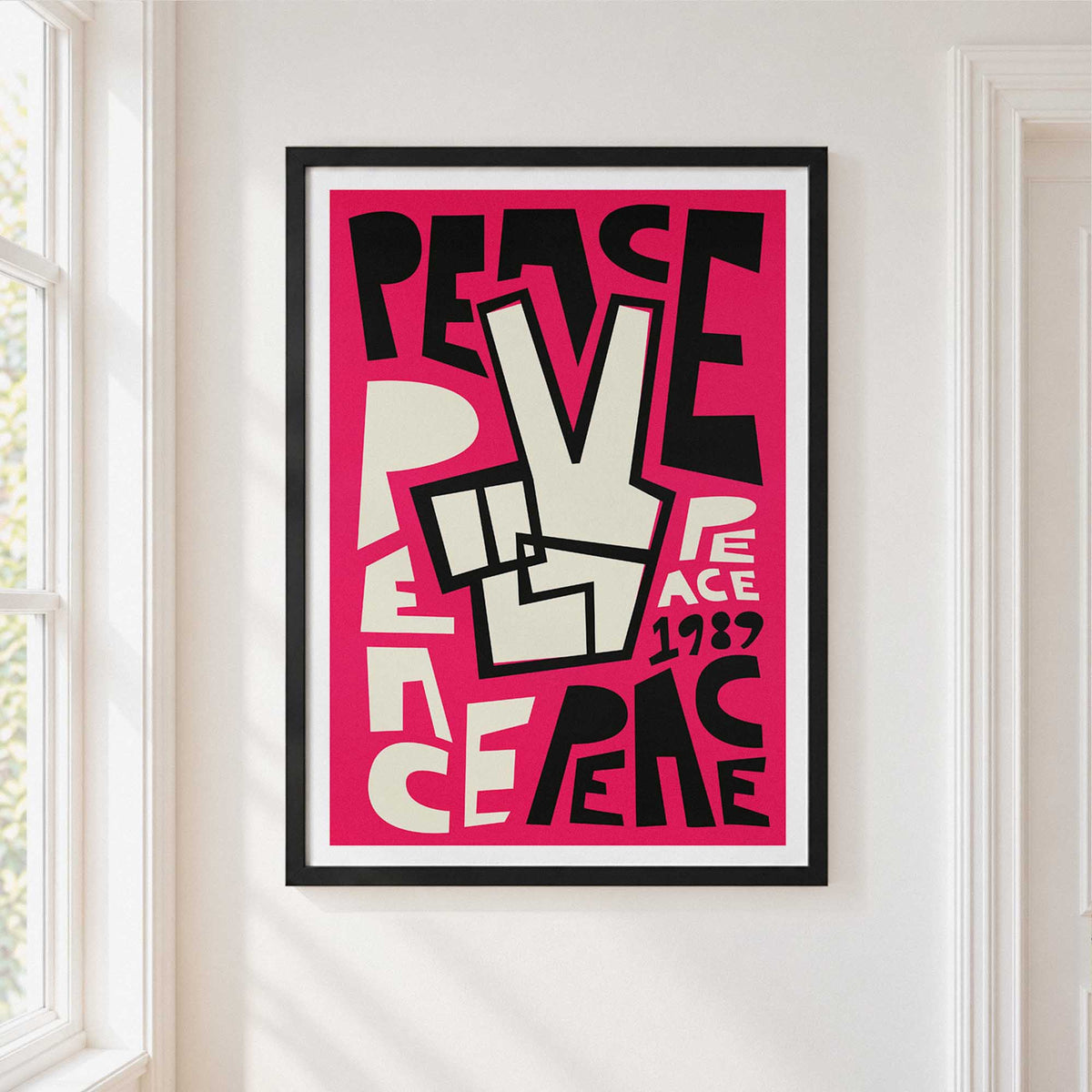 A framed art print featuring a bold pink, black, and off-white graphic with the lettering & typography 'PEACE' and a peace sign, hung on a white wall next to a window.