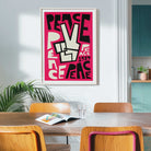 A framed print with a bold pink, black, and cream lettering and typography peace design hangs on a light gray wall in a dining room setting with wooden chairs and a table.