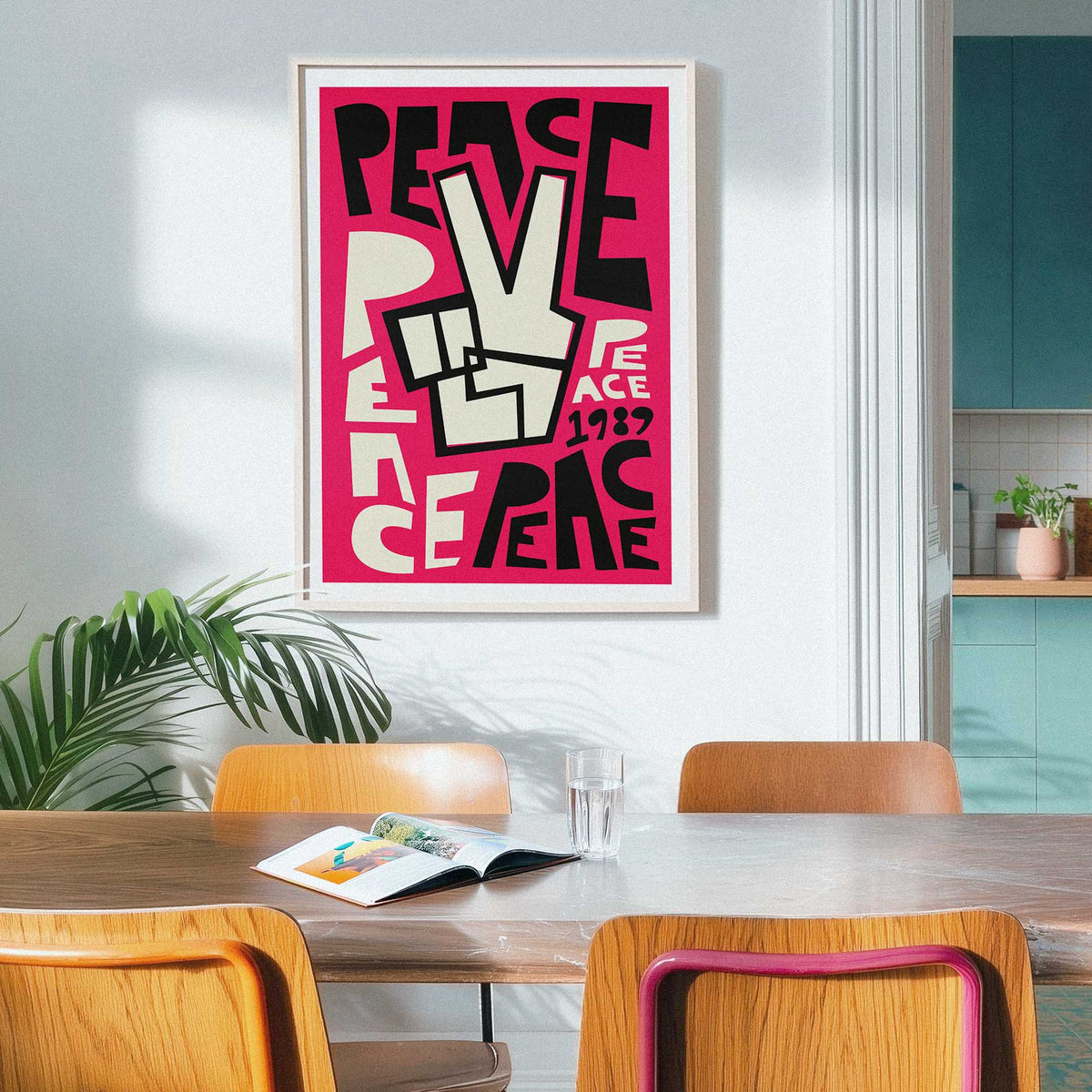A framed print with a bold pink, black, and cream lettering and typography peace design hangs on a light gray wall in a dining room setting with wooden chairs and a table.