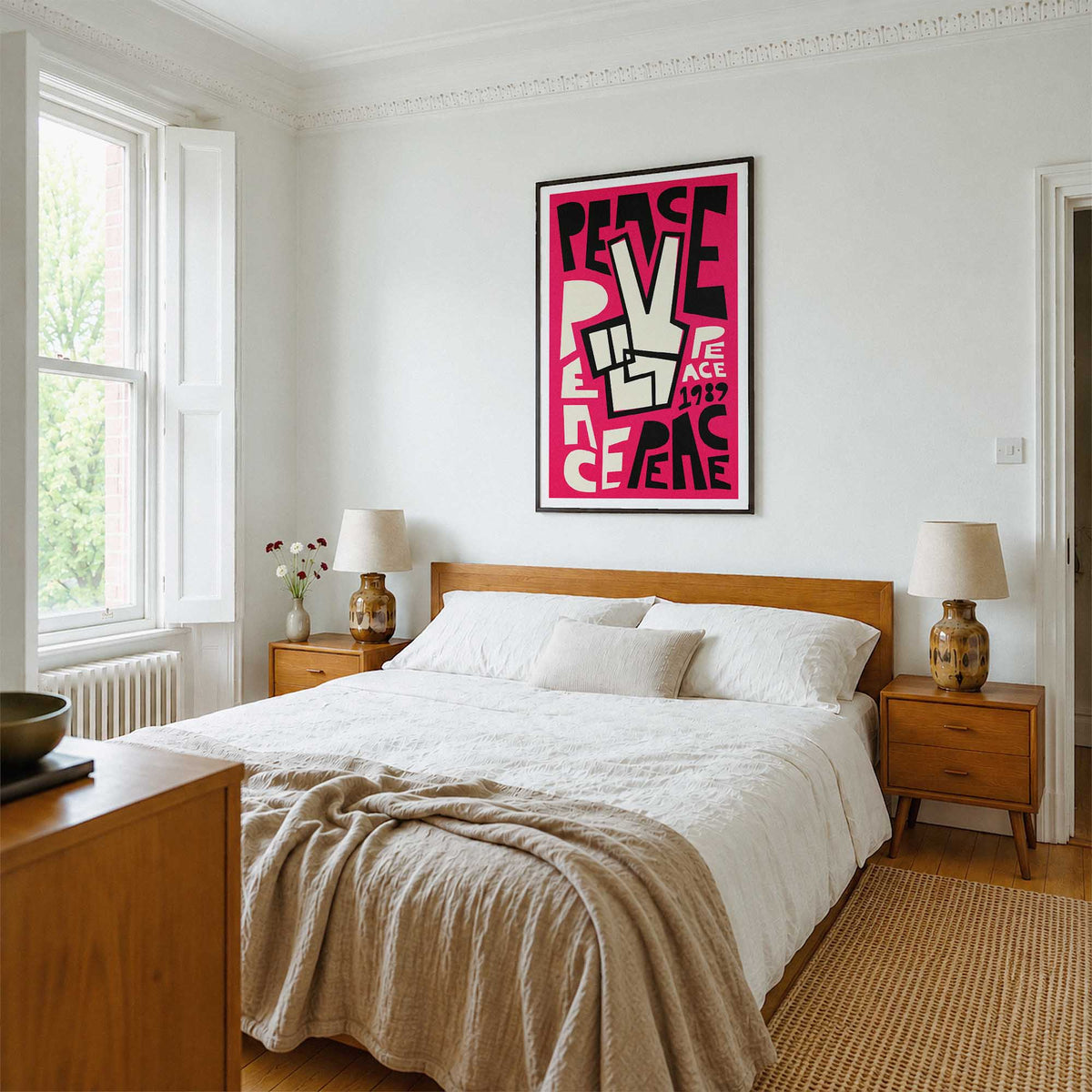 A styled bedroom interior featuring a framed graphic art print with the lettering and typography 'PEACE' in bold pink and black lettering, with a white peace sign. The print is mounted on a white wall above a wooden bed with white linens and a beige throw blanket.