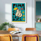 A framed art print featuring a colorful cocktail glass illustration with the text 'PIÑA COLADA' hangs on a light gray wall in a modern dining room setting with wooden chairs and a table.