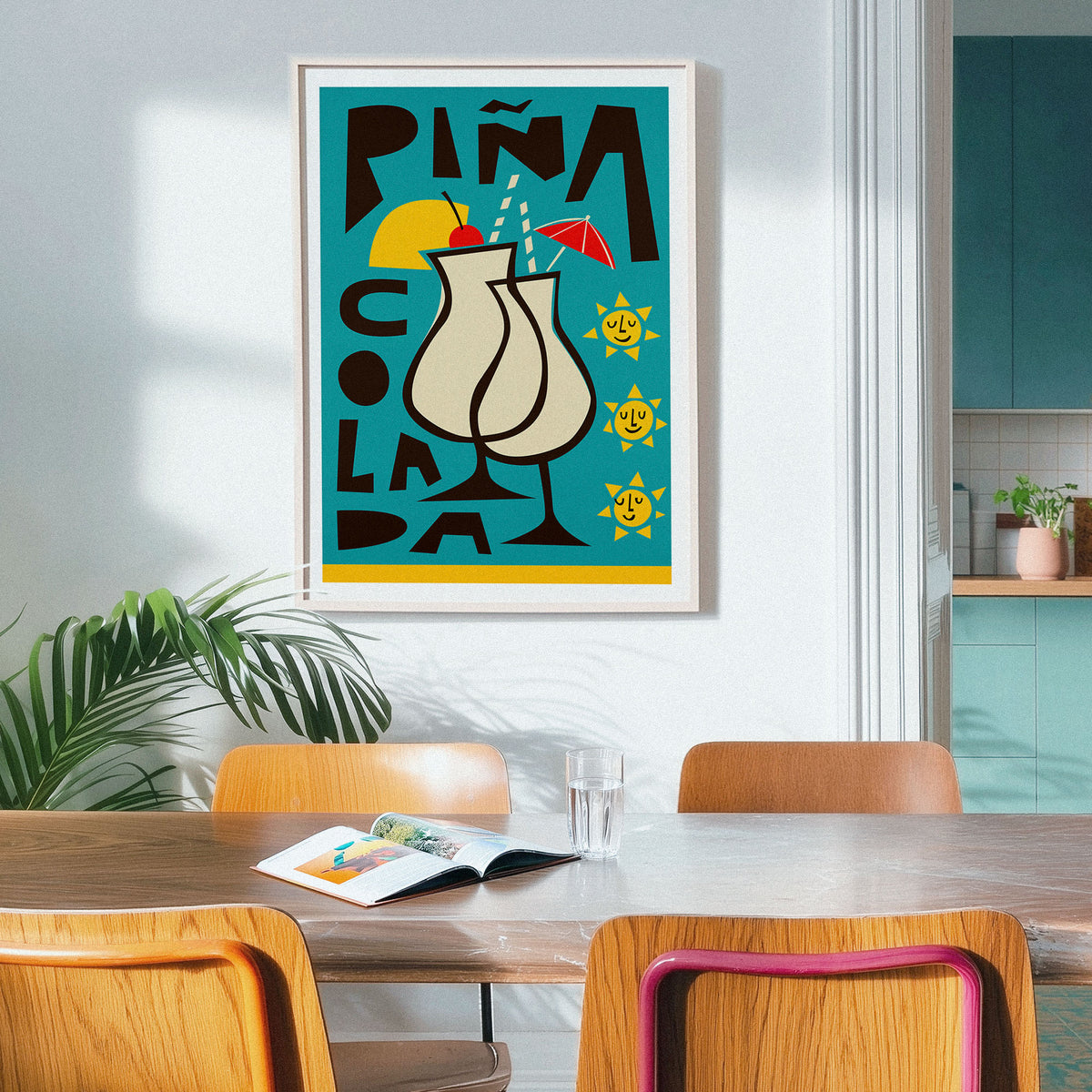 A framed art print featuring a colorful cocktail glass illustration with the text 'PIÑA COLADA' hangs on a light gray wall in a modern dining room setting with wooden chairs and a table.