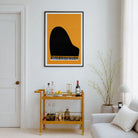 A framed art print featuring a minimalist black grand piano silhouette on a mustard yellow background, displayed on a white wall in a modern living room setting above a gold bar cart.
