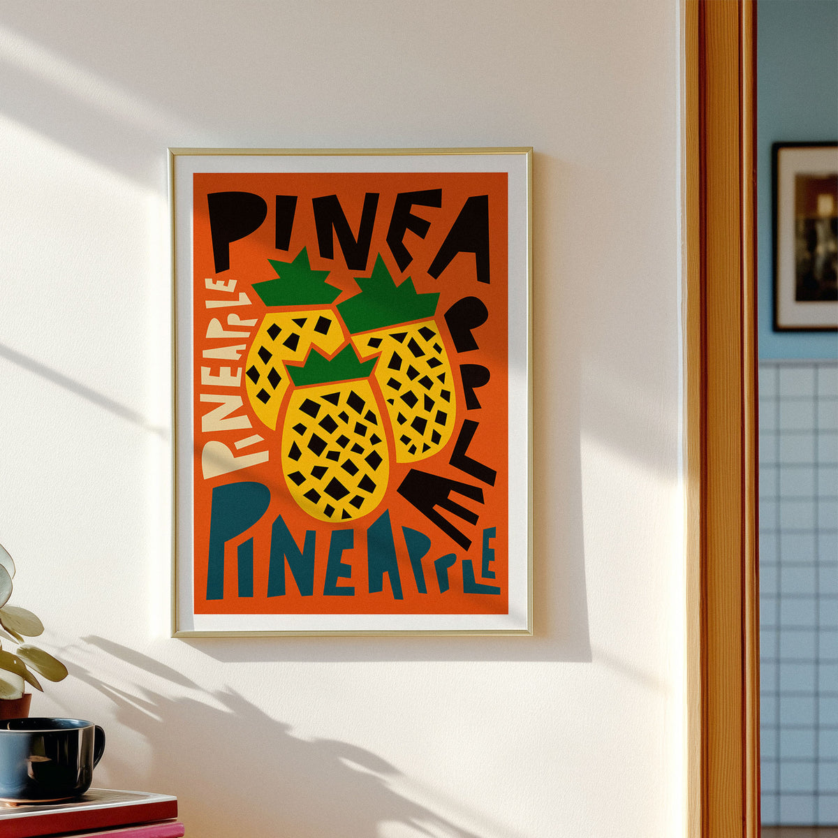 A framed art print featuring a retro-style illustration of three pineapples on an orange background with bold black and blue text, hanging on a white wall in a sunlit room.