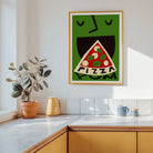A framed art print featuring a stylized green face eating a slice of pizza with the word 'PIZZA' inside, hanging on a white kitchen wall above a counter with a plant and mugs.