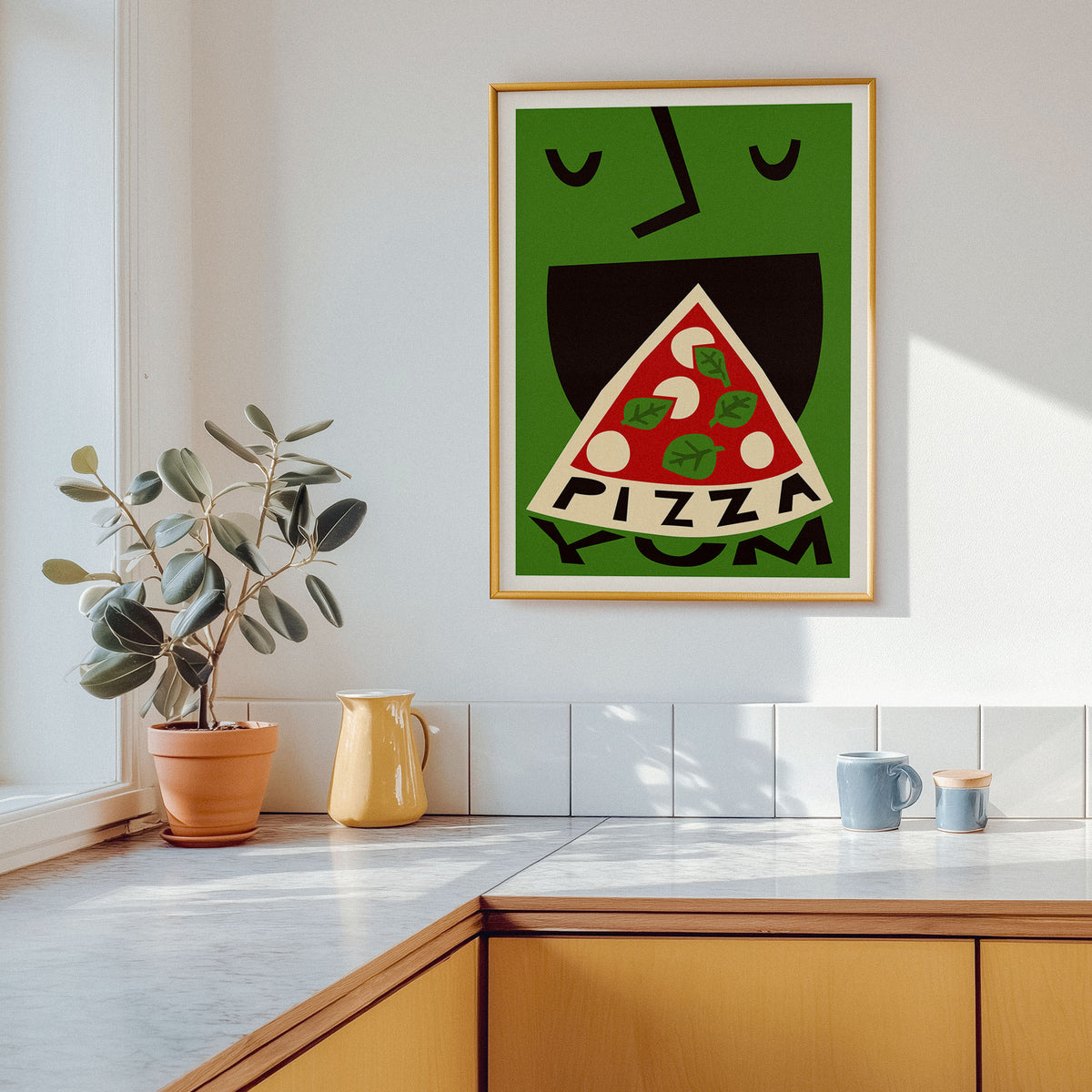 A framed art print featuring a stylized green face eating a slice of pizza with the word 'PIZZA' inside, hanging on a white kitchen wall above a counter with a plant and mugs.