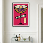 A framed pop-art style print of a martini cocktail with the word 'martini' on the glass, hanging on a white wall above a bar cart with bottles and glasses.