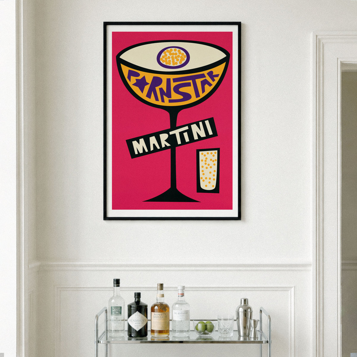 A framed pop-art style print of a martini cocktail with the word 'martini' on the glass, hanging on a white wall above a bar cart with bottles and glasses.
