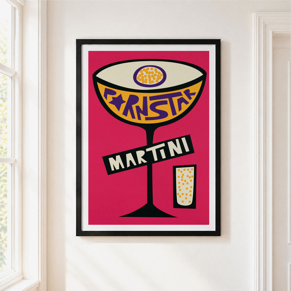 Framed pop art print of a martini cocktail on a pink background, hanging on a white wall next to a window.